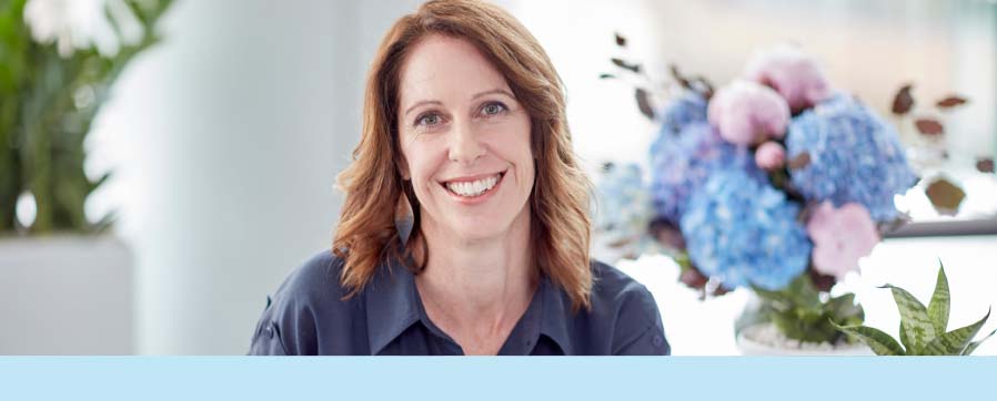 A picture of a lady smiling ay the camera. There are flowers behind her head.